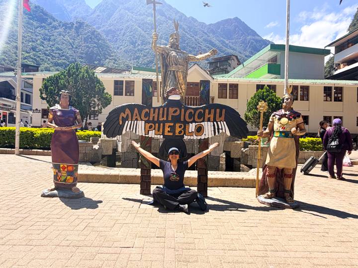 Traveler poses with arms wide at the Machu Picchu Pueblo welcome statue ensemble in the town square.