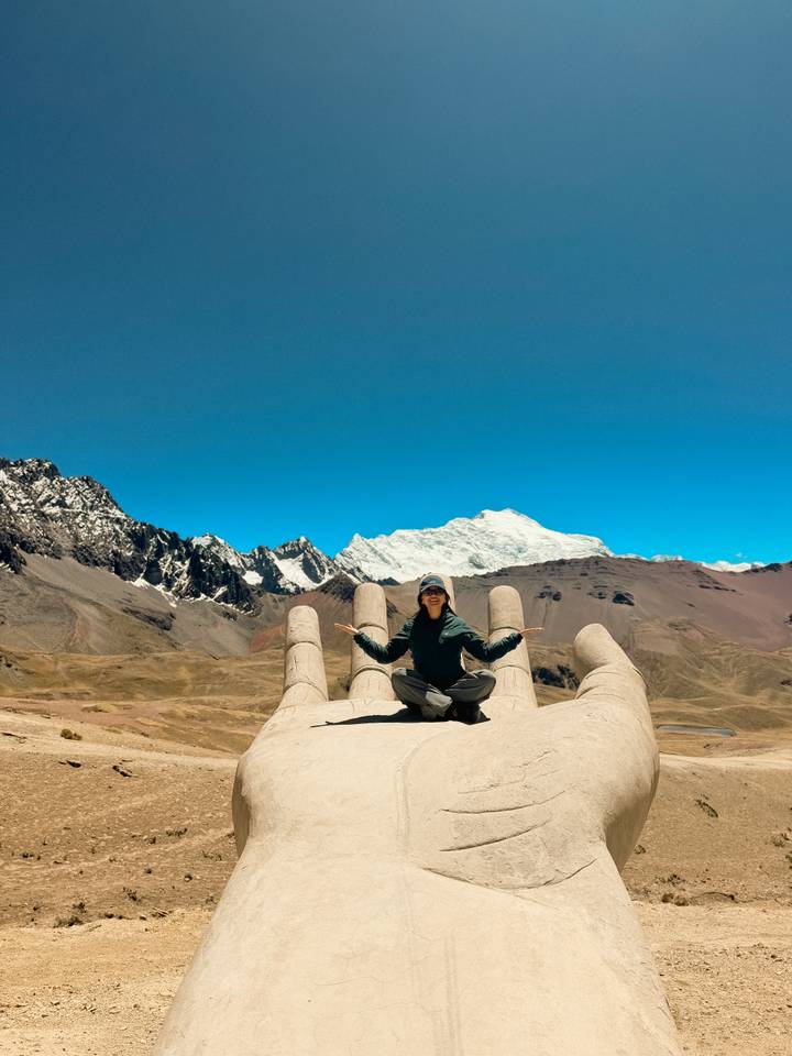 Traveler sits cross-legged on a giant concrete hand sculpture high in the Andes with glaciers beyond.