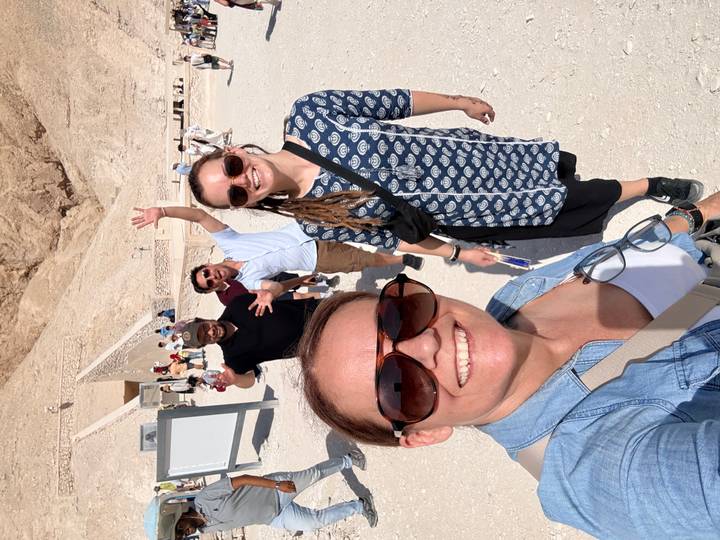Five smiling travellers pose in bright sunlight on a sandy pathway near ancient stone tombs and cliffs.