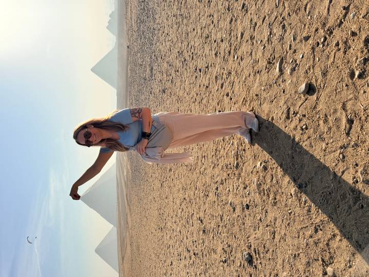 Tourist playfully poses as if pinching the top of a distant pyramid on a sandy plain under bright sun.