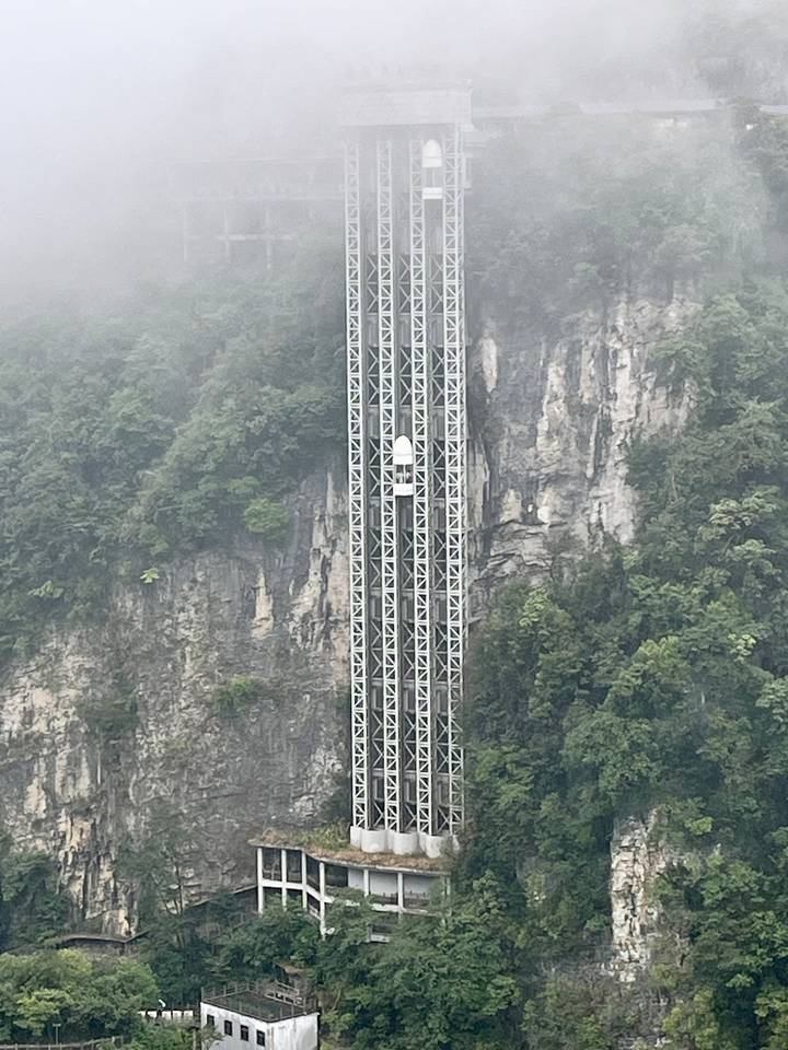Tall glass-fronted elevator shaft rises against a misty cliff face covered in greenery.