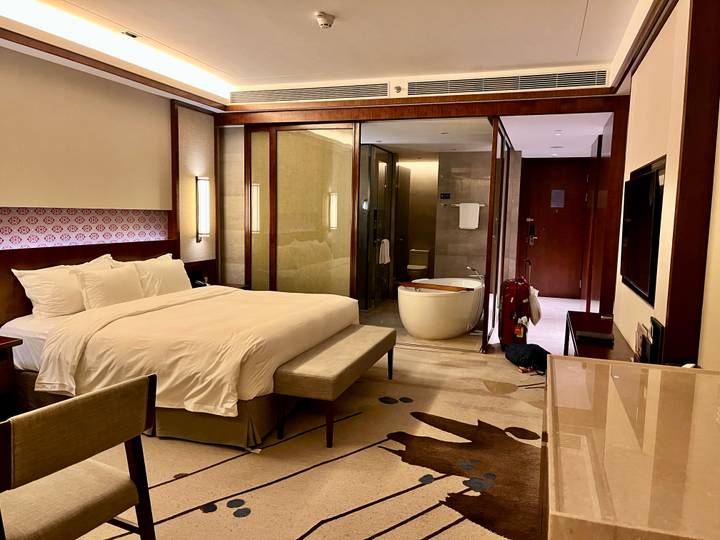 Modern hotel room with large bed, glass-walled bathroom and standalone soaking tub.