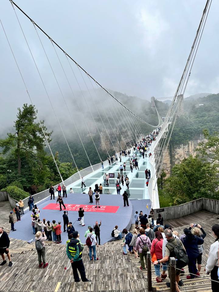 Crowds walk across a long glass suspension bridge spanning a misty canyon with forested cliffs.