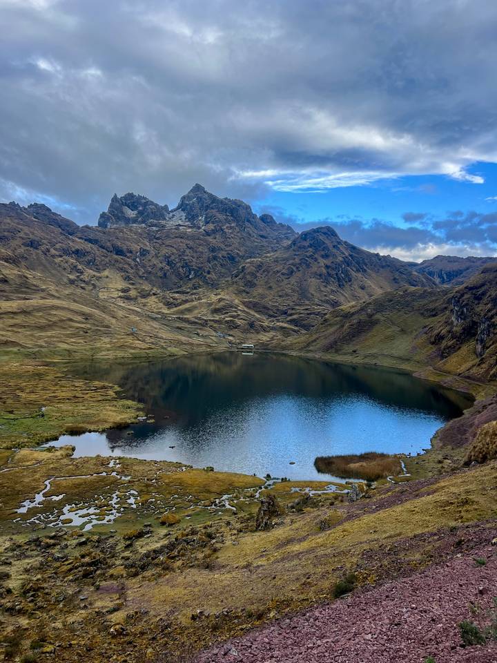 Serene mountain lake mirrors rugged Andean peaks and grassy golden slopes under a blue sky.