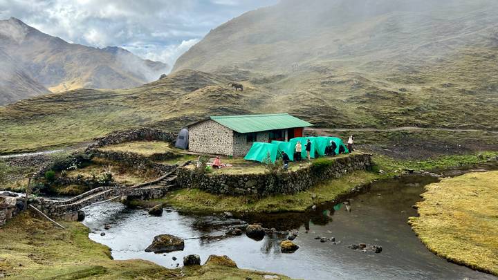 Small riverside campsite with green tents beside a stone hut and misty Andean hills.