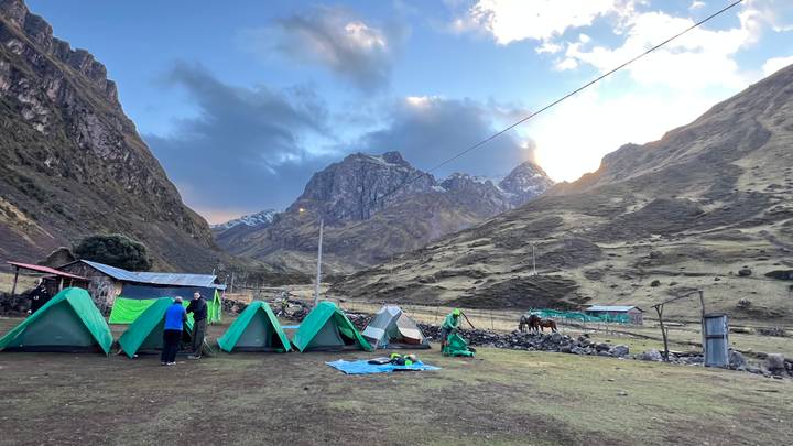 Late-afternoon campsite with green tents set beneath towering snowy peaks as the sun dips behind mountains.