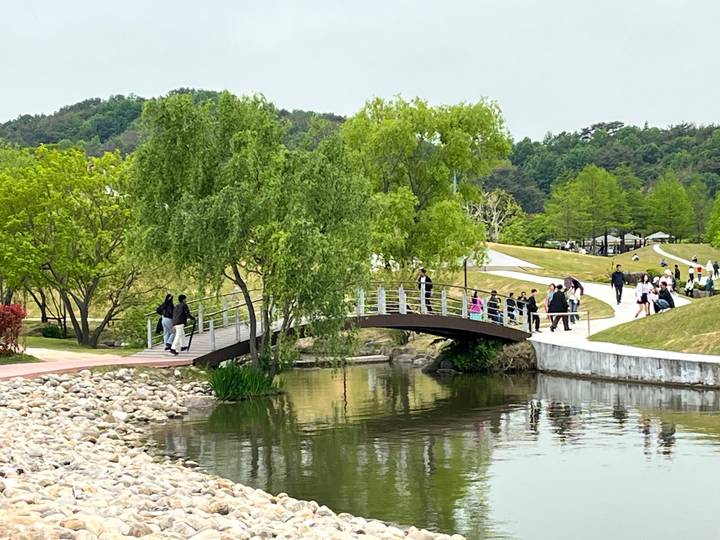 Park visitors stroll across a small arched bridge over a reflective pond surrounded by fresh spring greenery.