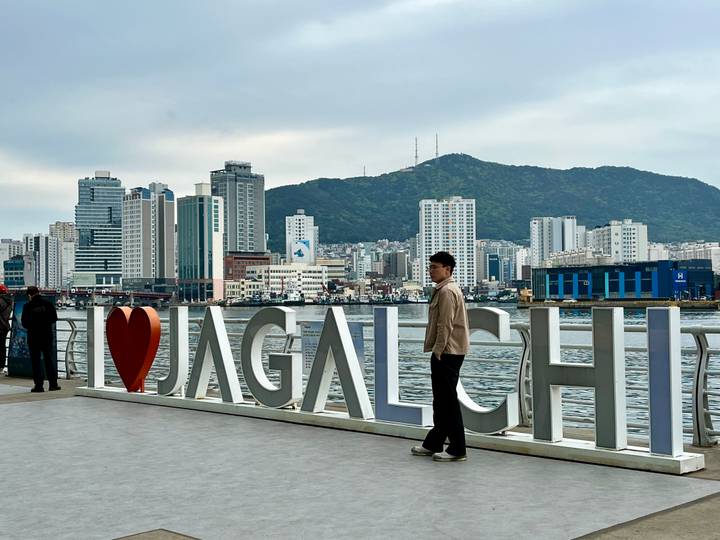 People pose beside the large waterfront 'I ♥ Jagalchi' sign with Busan skyline and mountain backdrop.