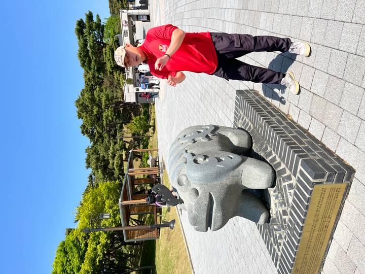 Guide gestures beside a carved stone bear statue in a park on a bright day.