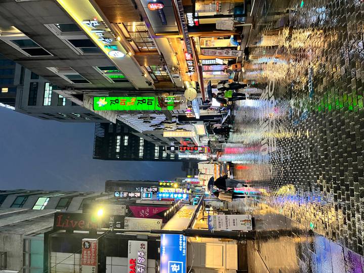 Rain-soaked neon shopping street at night with reflections glistening on cobblestones and people under umbrellas.