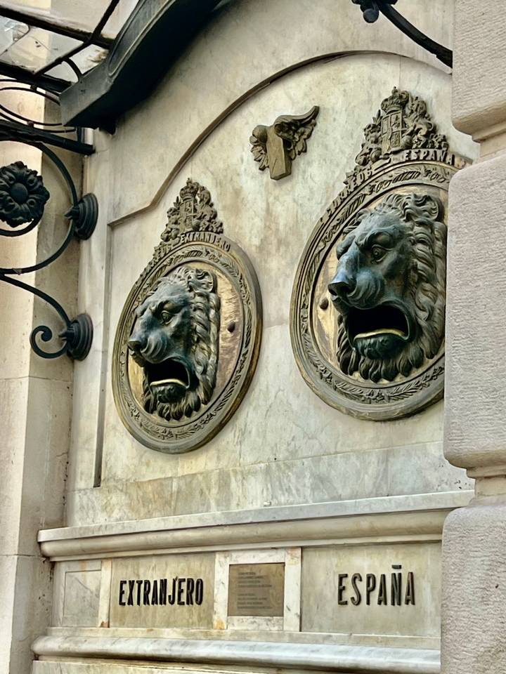 Pair of bronze lion-head mail slots mounted on a marble wall, mouths open wide.