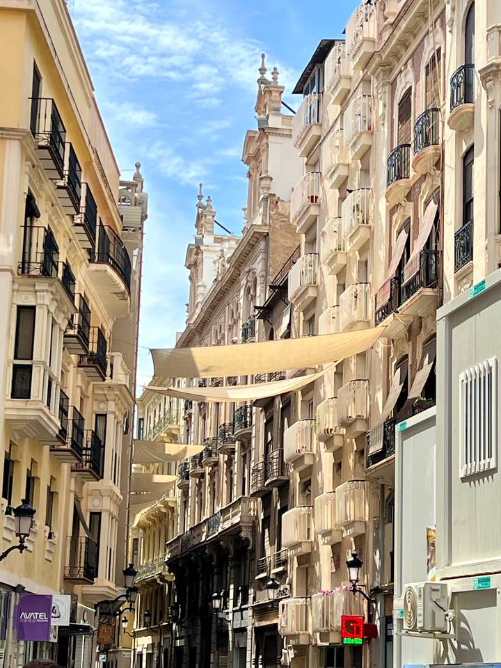 Narrow Spanish street lined with balconies and beige buildings shaded by stretched fabric awnings.