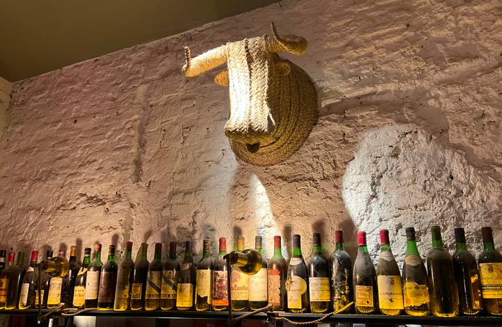 Rustic bar wall with rows of vintage wine bottles beneath a woven straw bull’s head mount.