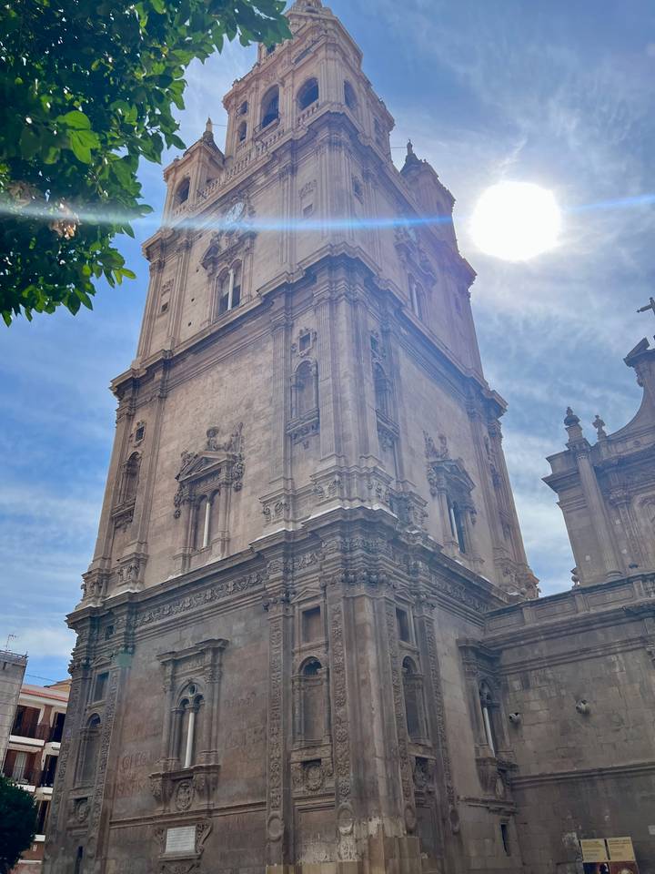 Ornate stone cathedral tower rises into a partly cloudy sky, seen from ground level.
