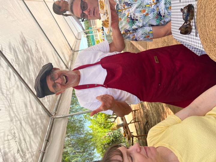 Smiling chef in red apron and black beret greeting guests at an outdoor dining setup under a canopy.