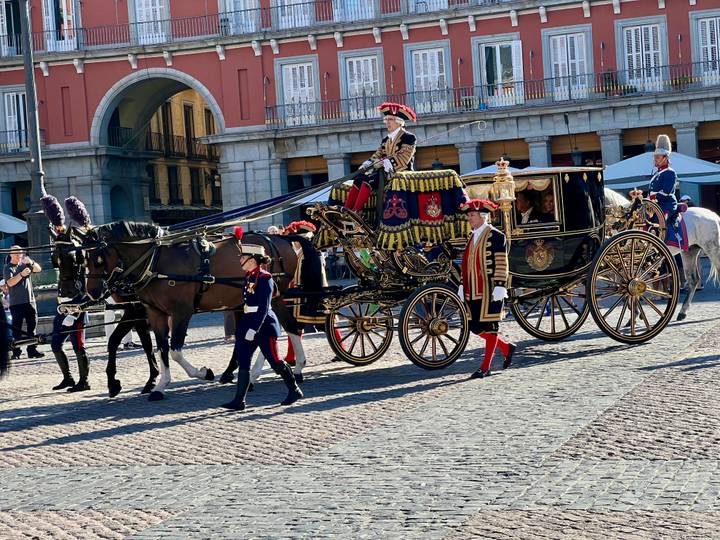 Ornate horse-drawn royal carriage escorted by guards crossing a historic plaza.