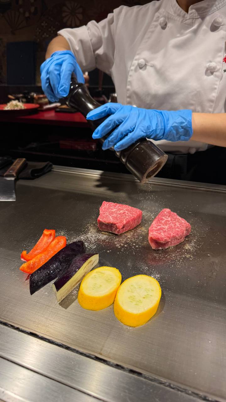 Chef seasons raw wagyu steaks on a teppanyaki grill alongside colorful sliced vegetables.