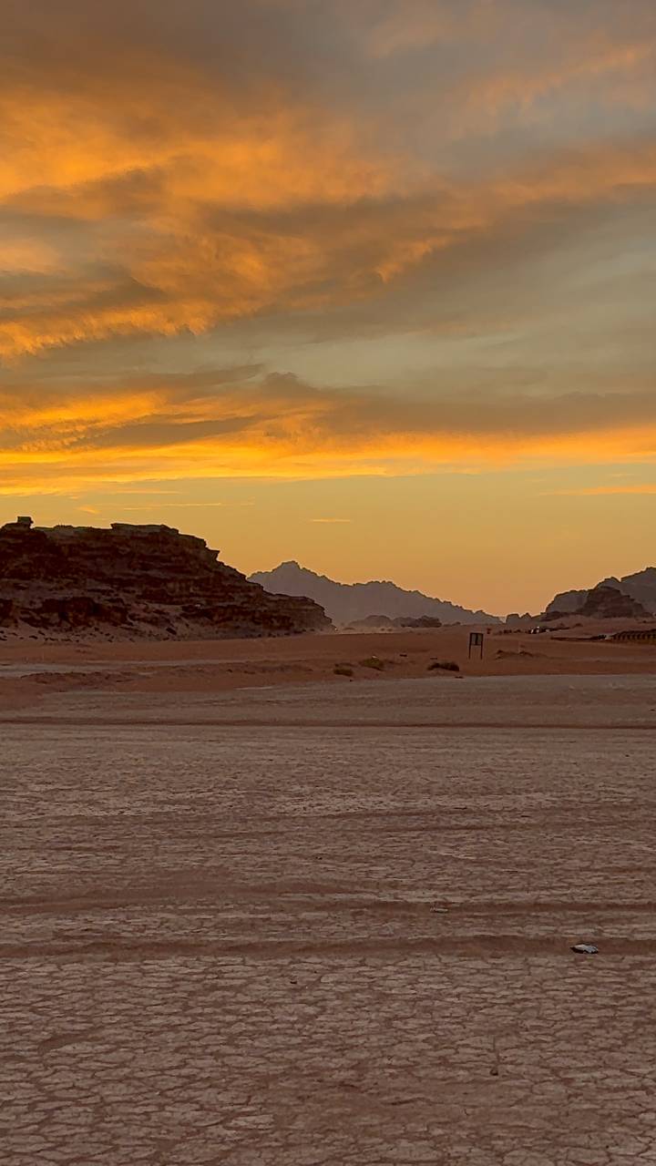 Golden desert landscape and distant jagged mountains illuminated by a vivid sunset sky in Wadi Rum.