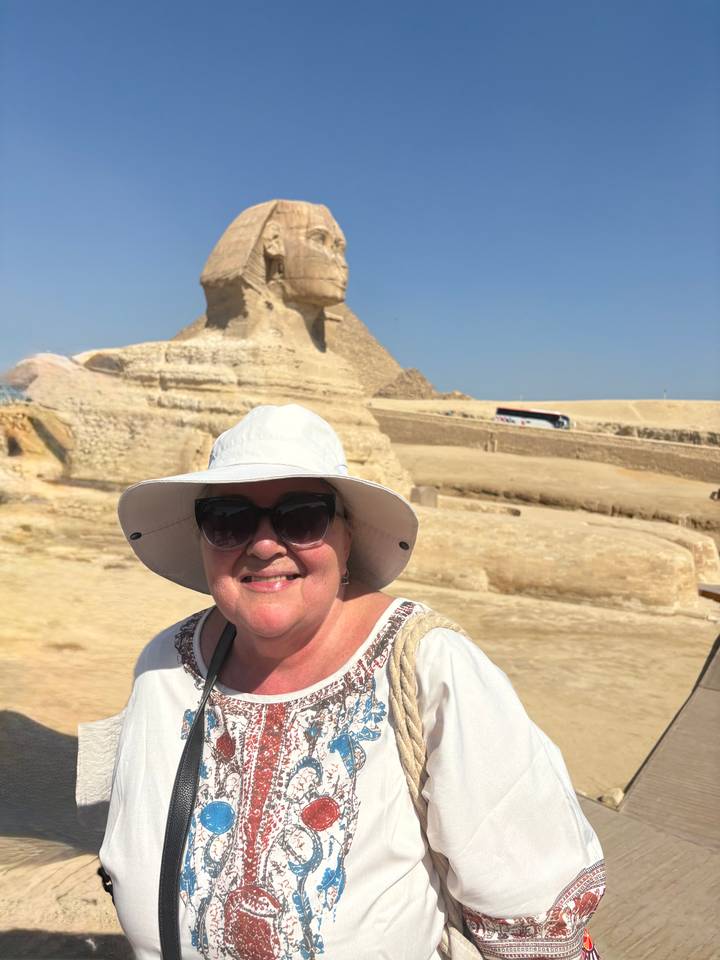 Smiling woman wearing a sun hat and sunglasses in front of the Great Sphinx under a clear blue sky.