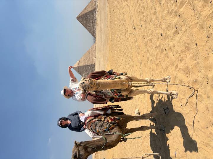 Two tourists riding camels on the sandy plateau with the Giza pyramids rising in the background.