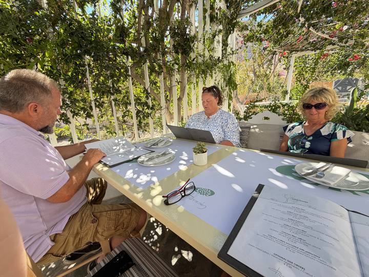 Three travelers reading menus at a shaded outdoor cafe with leafy backdrop.