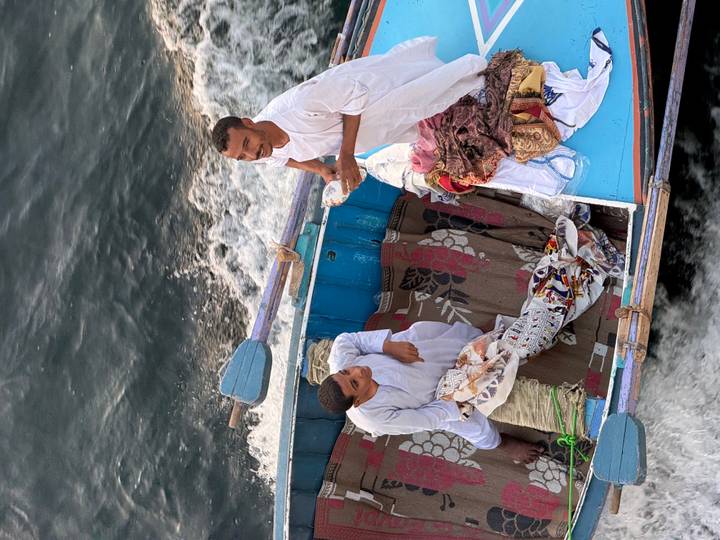 Two men in white robes ride a small wooden boat laden with rugs on swift river water.