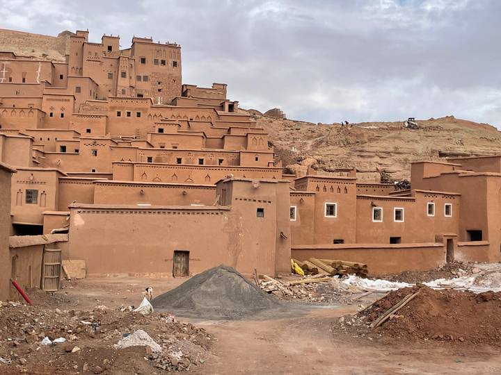 Earthen adobe village of Ait Benhaddou with terraced houses climbing a hillside under cloudy sky.
