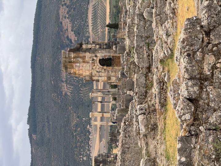 Ancient stone ruins and columns of a Roman archway stand among scattered blocks with green hills behind.