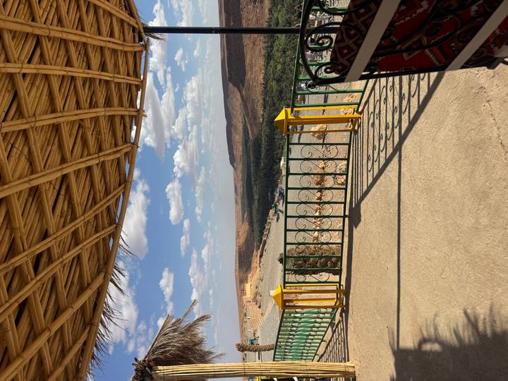 Terrace with bamboo roof frames a sweeping desert canyon and distant palm oasis under a blue sky.