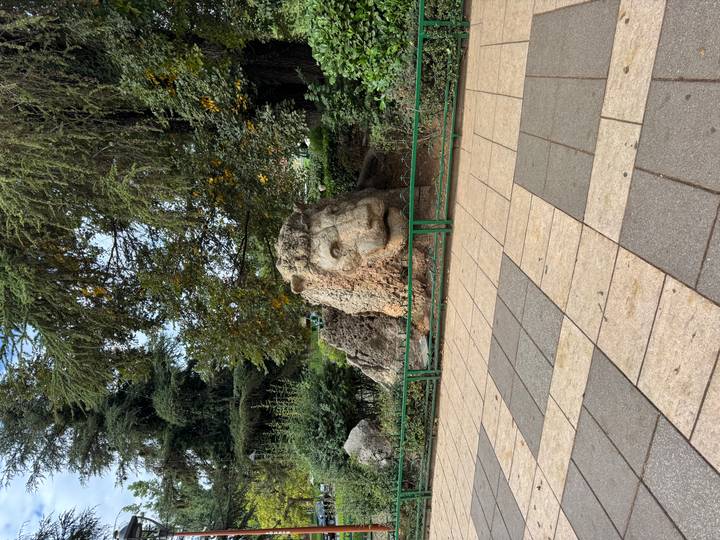 Large carved stone lion head sculpture sits among greenery beside a tiled walkway in a park.