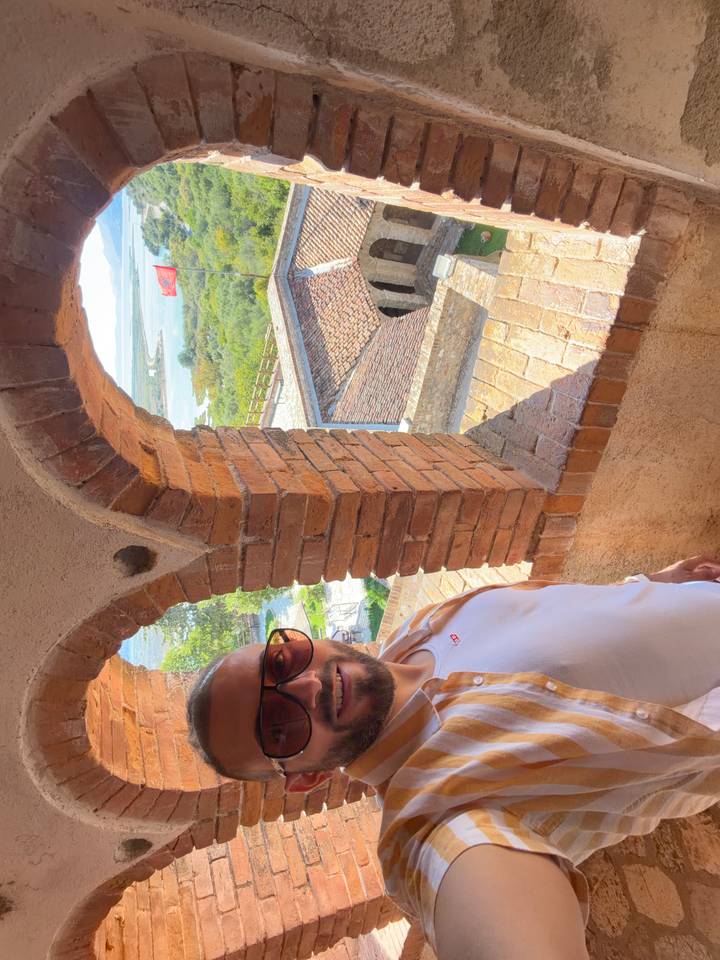 Man in sunglasses poses inside a brick-arched tower window overlooking greenery and rooftops.