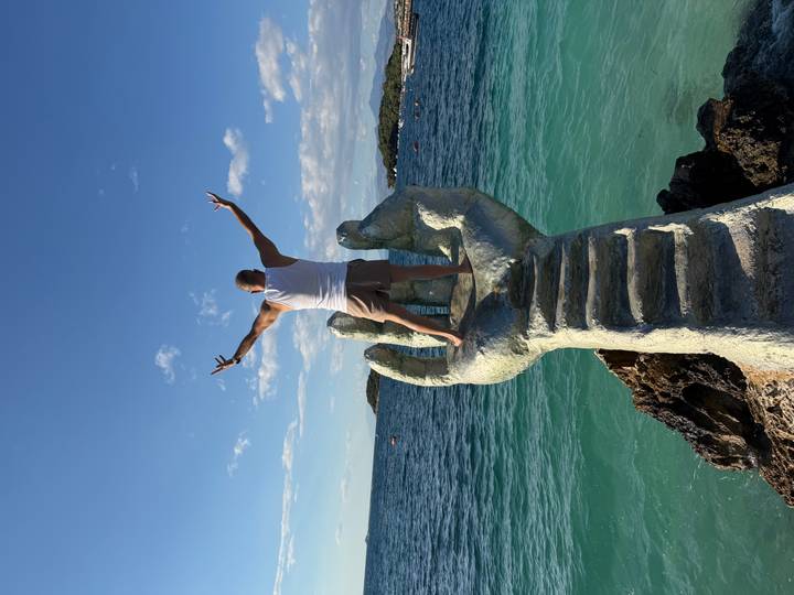 Victorious man raises arms atop a giant hand statue emerging from turquoise sea under blue sky.