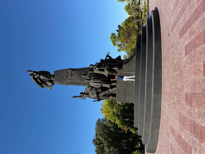 Visitor stands on the steps of a monumental bronze statue complex reaching into a clear blue sky.