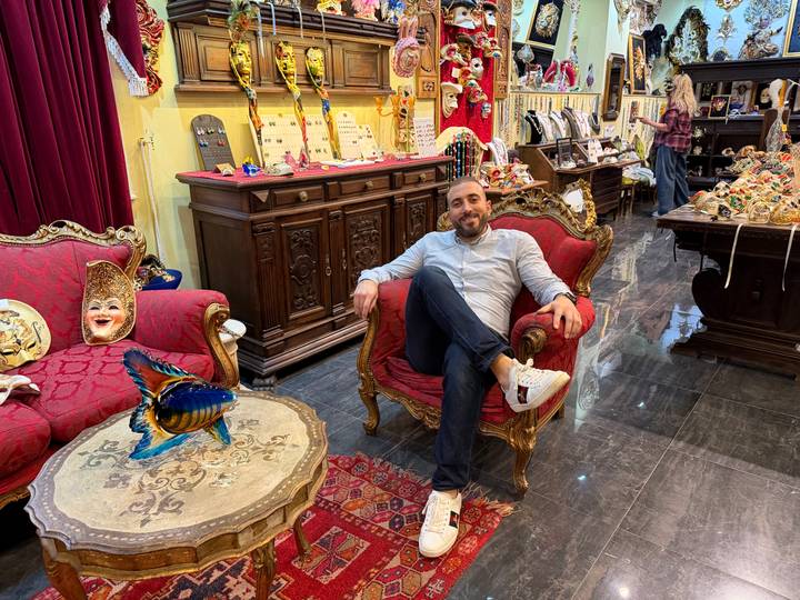Man lounges on an ornate red armchair inside a lavish curio shop filled with colorful masks and trinkets.