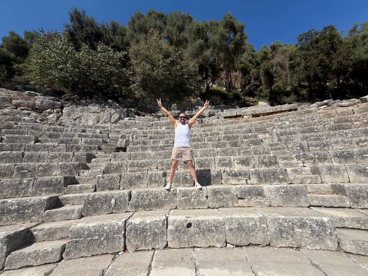 Traveller throws up peace signs while standing in the stone tiers of an ancient amphitheatre surrounded by trees.