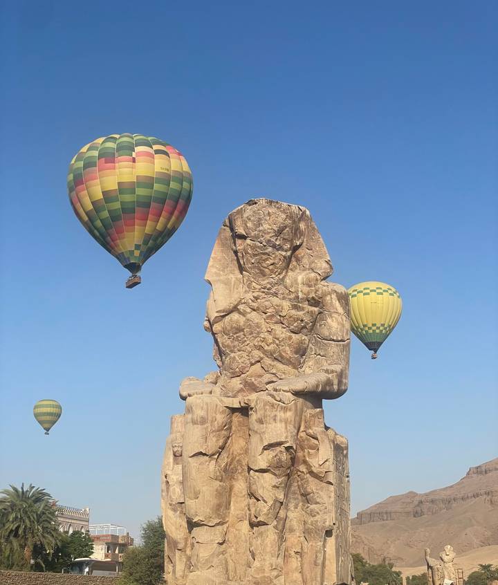 Hot-air balloons rise behind the weathered Colossus of Memnon against a crystal blue Luxor sky.