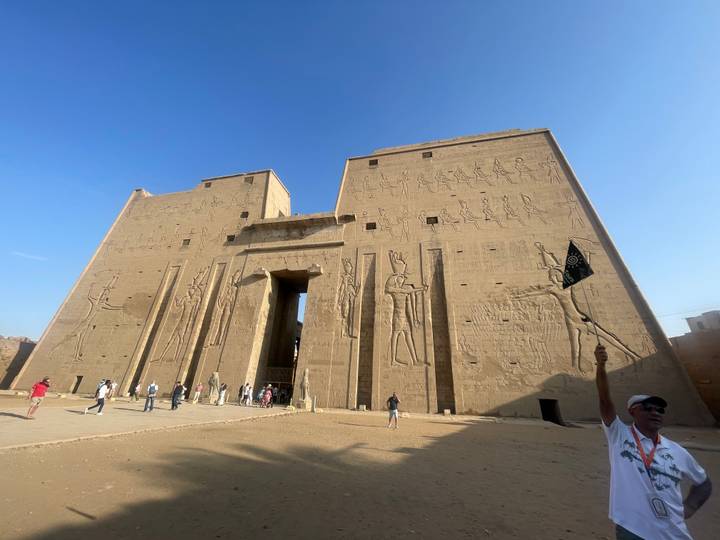 Grand sandstone façade of Edfu Temple covered in reliefs with tourists and guide holding flag for scale.