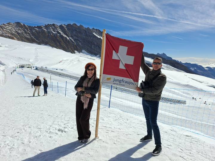 Two travellers stand on snowy plateau at Jungfraujoch holding a Swiss flag with dramatic alpine peaks behind.