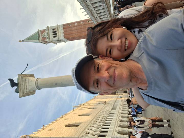 Selfie of couple in Piazza San Marco with the lion-topped column and Campanile rising into a pale sky.