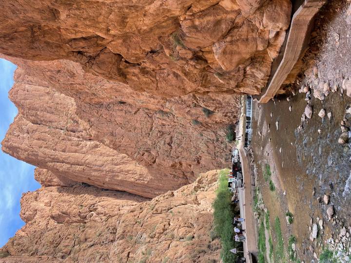 Towering red cliffs of Todra Gorge frame a stream and footpath with vehicles parked below.
