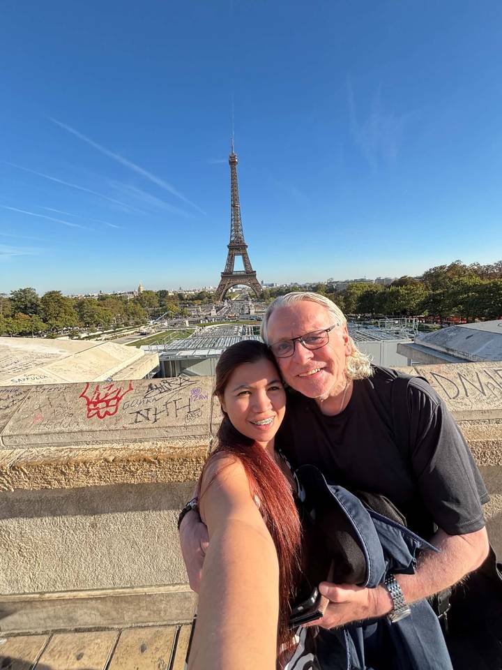 A smiling couple takes a selfie with the Eiffel Tower rising behind them on a clear sunny day.