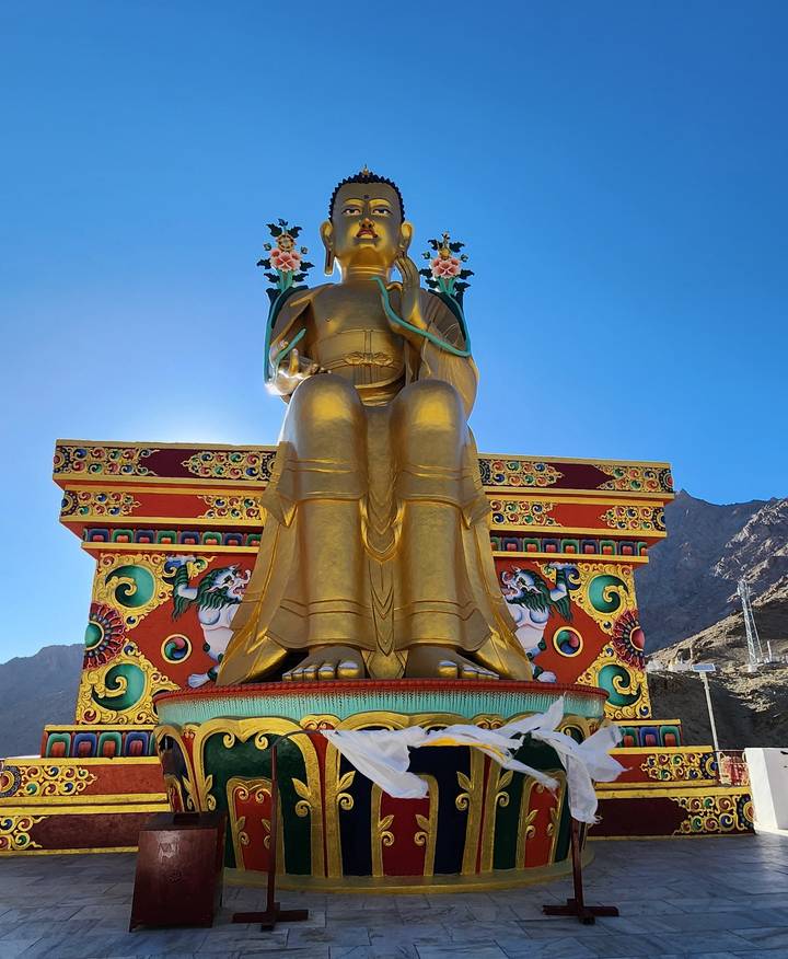 A giant golden seated Buddha statue, backlit by clear blue sky, stands on a vividly painted pedestal.