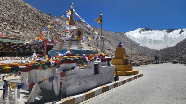 Prayer-flag-draped chorten stands beside a glaciated mountain pass road under a clear blue sky.