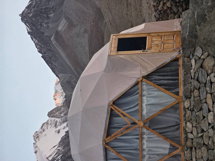 A geodesic dome tent with wooden door and curtains set against rugged grey mountains with a snow-tipped peak catching warm light.