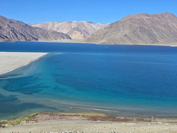 Vivid turquoise and deep-blue waters of a high-altitude lake contrast with pale sandy spits and arid mountains surrounding Pangong Tso.