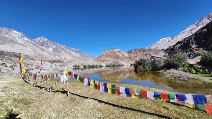Colourful prayer flags line a lakeside meadow with reflections of barren peaks and clear blue sky in still water.
