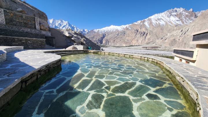 A stone-lined thermal pool with crystal-clear water overlooks a dramatic, snow-capped valley under bright blue skies.