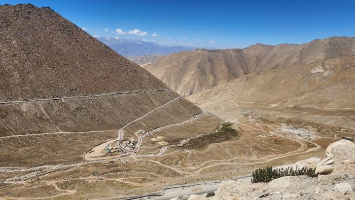 A winding high-altitude road snakes across a barren brown mountain landscape under clear skies.