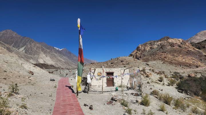 A small white shrine adorned with prayer flags stands in a stark rocky desert valley.