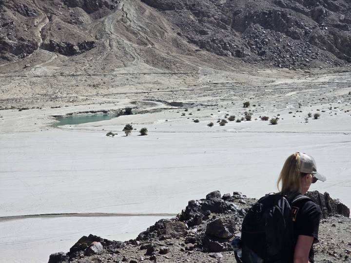 A lone hiker with a backpack looks out over a wide pale riverbed and rocky desert landscape.